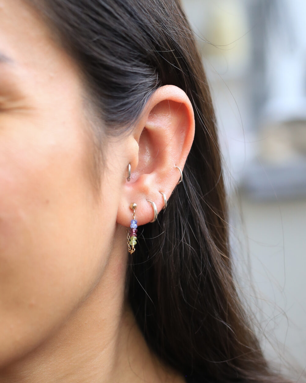 Het model draagt korte edelsteen oorhangers van roze en blauwe saffier en lichtgroene peridoot edelsteen. Afgewerkt met gouden oorsteker en kort kettnkje